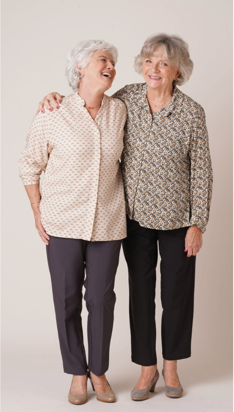 Deux femmes seniors souriantes se tiennent par les épaules. Elles portent des chemisiers à motifs et des pantalons élégants, posant côte à côte dans une ambiance chaleureuse et bienveillante - forESTIME